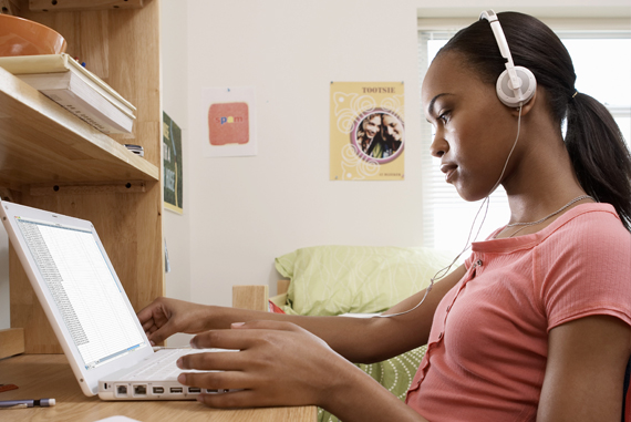 A female African-American student sits at a desk in her dorm room, wearing headphones and working on a laptop computer.