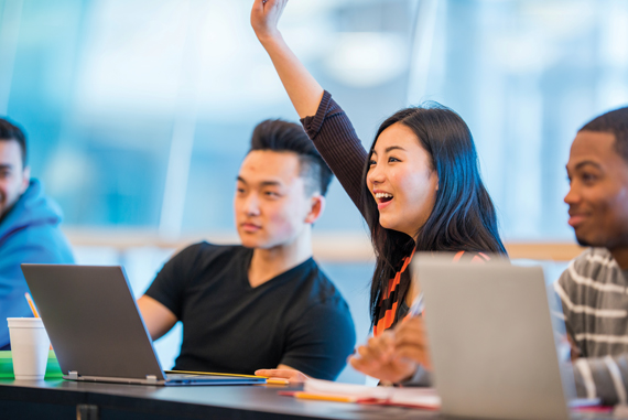 Asian college student raising her hand in class, while seated at a long table with other students. 